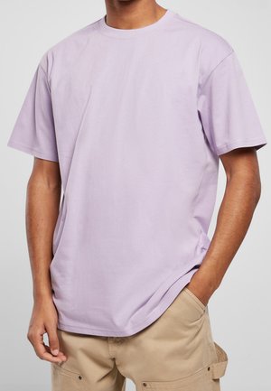 Person iført en lys lilla T-shirt med korte ærmer og beige cargobukser med den ene hånd i lommen mod en ensfarvet baggrund.