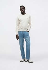 Ανοιχτό μπεζ φούτερ με ριμπ μανσέτες, ανοιχτό μπλε τζιν παντελόνι με ίσιο κόψιμο και γκρι και λευκά αθλητικά παπούτσια με μαύρες ρίγες. Απλός, μινιμαλιστικός σχεδιασμός.