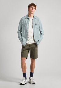 Camicia di jeans blu chiaro, t-shirt bianca, pantaloni corti verde oliva, sneakers grigie con dettagli bianchi, calze blu navy, in piedi contro uno sfondo neutro.