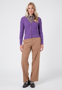 Lila kabelstickad cardigan med knappar, i kombination med en mönstrad skjorta och bruna byxor. Skorna har dekorativa detaljer.