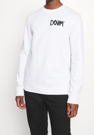 Hombre vistiendo sudadera de manga larga blanca con el texto "DENIM" en el pecho y pantalones negros, de pie frente a un fondo ligero y uniforme.
