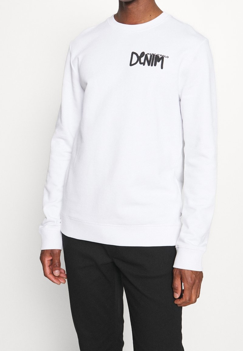 Man som bär en vit långärmad sweatshirt med texten "DENIM" på bröstet och svarta byxor, stående mot en plain ljus bakgrund.