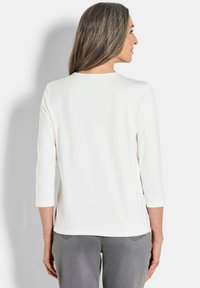 Weißes Oberteil mit drei Viertelärmeln, rundem Halsausschnitt und glatter Textur. Kombiniert mit grauen Jeans, die eine entspannte Passform und ein einfaches Design zeigen.