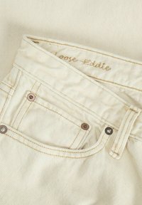 Traperice boje kreme s visokim strukom, s bakrenim kopčama, malim prednjim džepom i prošivenim detaljima na pojasu.