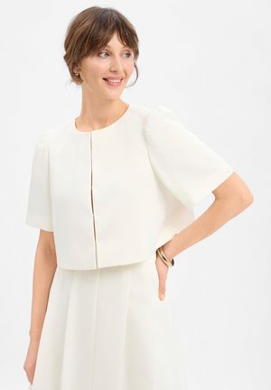 Vrouw met kort bruin haar draagt een wit cropped jasje met korte mouwen over een bijpassende jurk, glimlacht met hand op heup tegen een witte achtergrond.