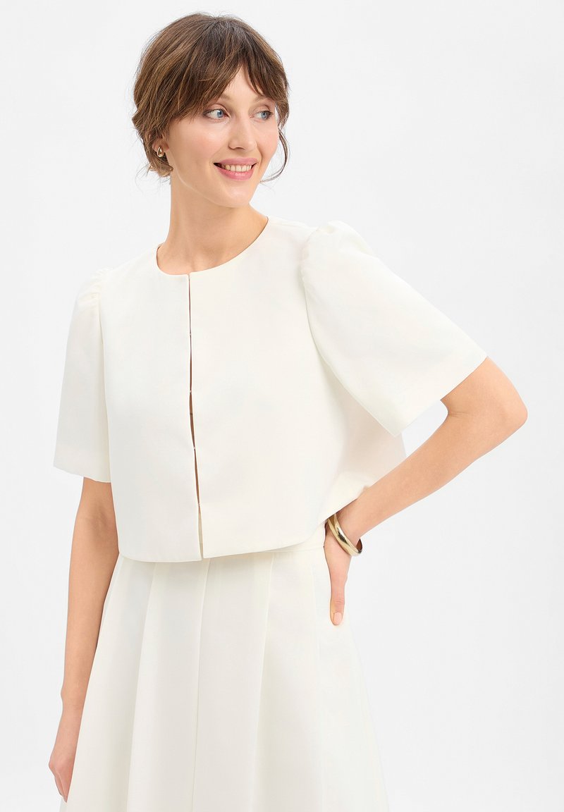 Frau mit kurzen braunen Haaren trägt eine weiße, kurzärmelige, bauchfreie Jacke über einem passenden Kleid, lächelt mit der Hand in der Hüfte vor einem weißen Hintergrund.