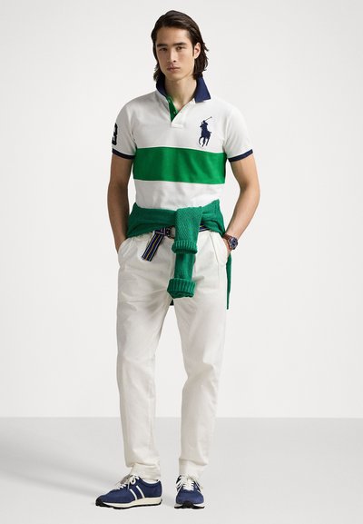 Balta polo krekls ar zaļām horizontālām svītrām, tumši zils apkakle un logotips. Sāna ar krēmkrāsas biksēm un tumši zilām kedām. Zaļš džemperis ap vidukli.