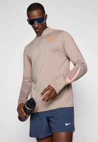 Top bej cu mânecă lungă, cu fermoar până la jumătate, logo portocalie și detalii roz pe mâneci, asortat cu pantaloni scurți bleumarin și o sticlă de apă neagră.