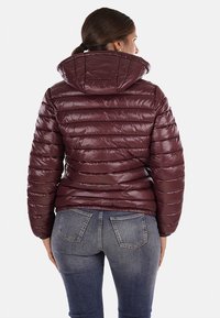 Giacca puffer bordeaux con cappuccio, caratterizzata da un design trapuntato e polsini elastici. Abbinata a jeans blu con tasche applicate.