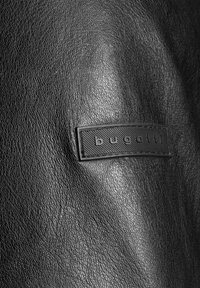 Zwarte leren stof met een getextureerd oppervlak. Bevat een rechthoekig zwart label met "bugatti" in reliëf en een gevlochten rand.