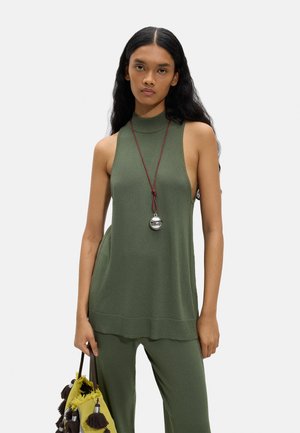 Kobieta w oliwkowej bluzce bez rękawów i spodniach, trzyma żółtą torbę z czarnymi frędzlami, nosi długi naszyjnik z okrągłym srebrnym wisiorkiem.