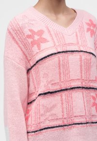 Personne portant un pull rose en tricot avec des motifs floraux, des rayures horizontales noires et un col en V.