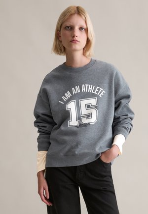 Grijze sweatshirt met witte tekst "I AM AN ATHLETE" en het nummer "15." Zachte textuur, ronde halslijn, afgekapte schouders, gelaagd met lichtcrème manchetten.