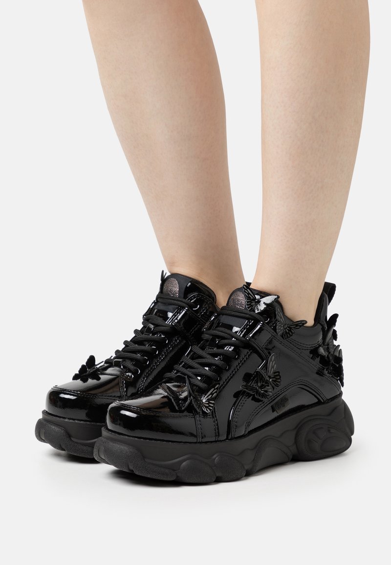 Black patent leather sneakers with a chunky sole, butterfly accents, and black laces. Features a padded collar and textured rubber outsole.