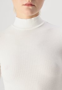 Hvit, ribbet halskjede topp laget av lett, elastisk materiale. Har høy halslinje og subtil tekstur med vertikale striper.