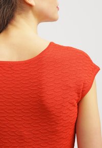 Röd texturerad topp med ett geometriskt mönster, breda axelband och korta ärmar. Tyget verkar slätt och lätt.