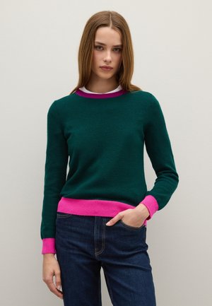 Grüner Wollpullover mit pinken Rippbündchen und -saum, Rundhalsausschnitt und langen Ärmeln, kombiniert mit dunkelblauen Jeans. Glatte Textur.