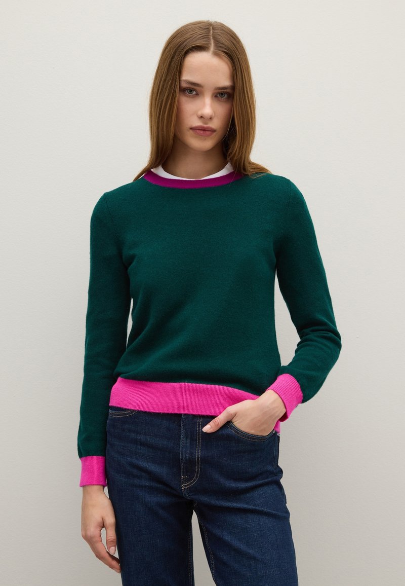 Maglione verde in lana con polsini e orlo rosa a coste, scollo rotondo e maniche lunghe, abbinato a jeans blu scuro. Texture liscia.