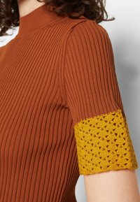 Ruskea nauhakudottu toppi, jossa on korkea kaula-aukko ja lyhyet hihat, sekä keltainen virkattu yksityiskohta hihansuun koristeena.