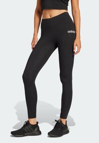 Svarta leggings i elastiskt material med hög midja och en liten vit Adidas-logotyp på vänster höft. Skorna är svarta.