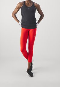Débardeur de sport noir et leggings rouges au design ajusté. Baskets noires avec des accents gris. L'athlète est debout dans une pose neutre.