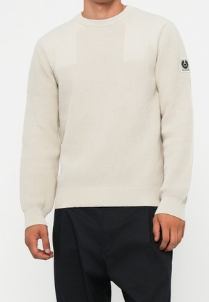 Hombre que lleva un suéter de punto texturizado color crema con cuello redondo y pantalones negros, con un parche con logo en la manga izquierda.