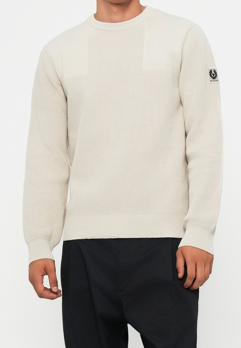 Homme portant un pull en maille texturée crème avec un col rond et un pantalon noir, avec un patch logo sur la manche gauche.