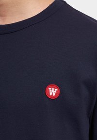 Granatowa bawełniana koszulka z okrągłym, czerwonym haftowanym logo z białą literą "W" na lewej części klatki piersiowej. Gładka, dopasowana tekstura.