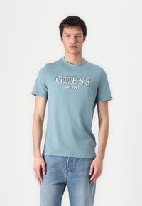 T-shirt in cotone azzurro chiaro con scollatura rotonda e maniche corte, con il logo "GUESS EST 1981" stampato in caratteri bold sul petto.