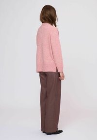 Roze gestructureerde trui met een hoge hals, gecombineerd met bruine geruite wijde broekspijpen en zwarte schoenen. Achteraanzicht, effen achtergrond.