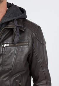 Ruskea nahkatakki, jossa on karhea rakenne, varustettuna vetoketjullisella rintataskulla, yksityiskohtaisella ompelulla ja tummanharmaalla huppusalustalla.