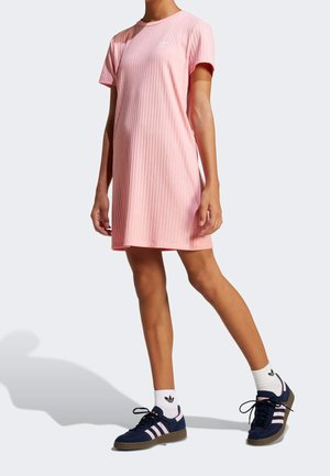 Kobieta modelka nosząca różową, prążkowaną sukienkę T-shirt z krótkimi rękawami oraz granatowe buty sportowe Adidas z różowymi paskami i białe skarpetki.