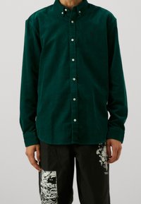 Mężczyzna w ciemnozielonej sztruksowej koszuli zapinanej na guziki oraz czarnych spodniach z białymi kwiatowymi nadrukami na udach na jednolitym tle.