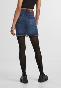 Denim mini-rok met een gefranjerde zoom, donkerblauwe wassing, achterzakken en een leren patch op de taille, gecombineerd met zwarte ondoorzichtige panty’s en stevige schoenen.