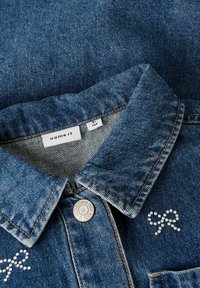 Džinsa jaka tumši zilā krāsā ar klasisku apkakli, metāla pogu un dekoratīvu spīdīgu rhinestone rakstu tauriņa formā.