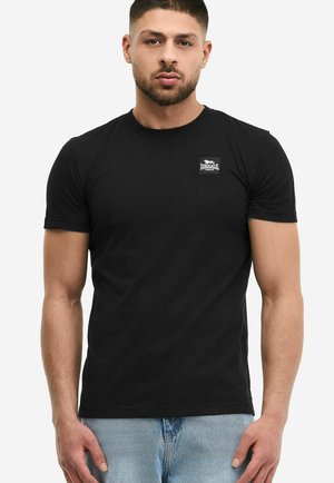 Ung mand med kort mørkt hår og skæg iført en tætsiddende sort Lonsdale London t-shirt og lyseblå jeans, stående foran en hvid baggrund.