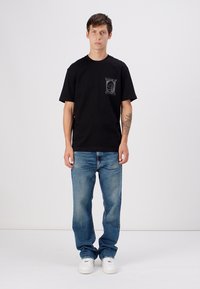 Black cotton t-shirt with a chest pocket featuring a print, paired with light blue denim jeans and white sneakers. Casual fit, crew neck.