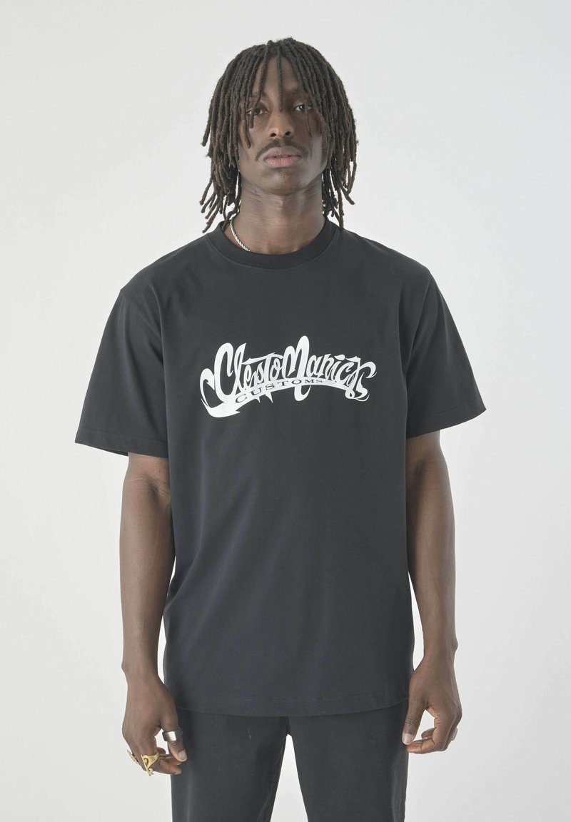 Mann mit schulterlangen Dreadlocks, der ein schwarzes T-Shirt mit weißem Aufdruck "Cletomanics Customs" trägt und mehrere Ringe an den Fingern hat.