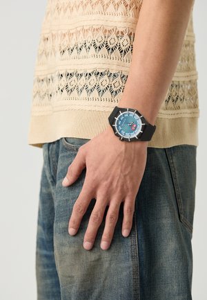 Montre bracelet noire avec un cadran rond bleu, accents métalliques et bracelet texturé, portée sur une main reposant contre un jean en denim bleu.