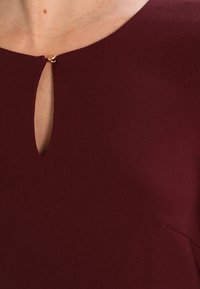 Burgundské šaty se výstřihem ve tvaru klíče se zlatým akcentem, vyrobené z hladkého materiálu s elegantním drapérií. Minimalistický design s čistými liniemi.