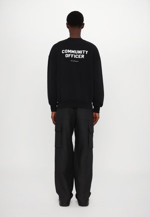 Svart sweatshirt med vit text "COMMUNITY OFFICER" på ryggen; bärs med lösa svarta cargobyxor och lågt profilskor.
