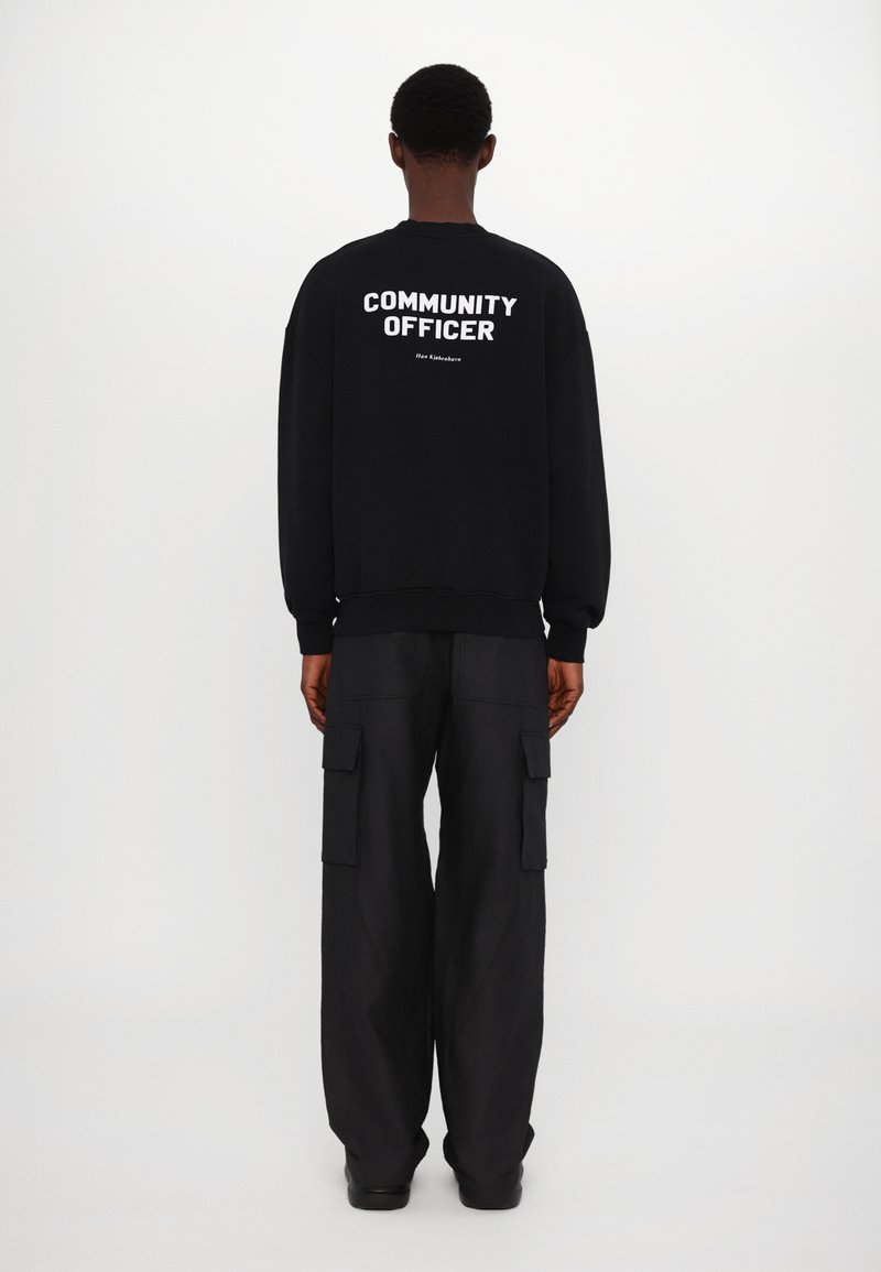 Sweat-shirt noir avec le texte blanc "OFFICIER DE COMMUNAUTÉ" dans le dos ; porté avec un pantalon cargo noir ample et des chaussures basses.