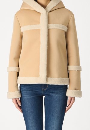 Veste en shearling beige avec bordure en peluche sur la capuche, les poignets, l'ourlet et la poitrine, portée avec un jean denim bleu, vue de face.