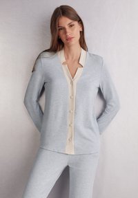 Helles blaues gestricktes Cardigan mit langen Ärmeln, cremefarbener Knopfleiste und rundem Kragen, kombiniert mit passenden Hosen. Glatte Textur.