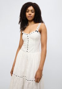 Λευκό φόρεμα από βαμβάκι με λουλουδάτη υφή με κεντήματα, κλείσιμο με κουμπιά, μαύρες λεπτομέρειες στους ιμάντες και το τελείωμα, εφαρμοστή μπούστα, αέρινη φούστα.