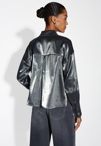 Metallic zilveren langemouwen shirt met een relaxte pasvorm, voorzien van een rugplooitje, gestructureerde kraag en knoopmanchetten, gecombineerd met donkere denim.