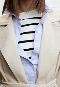 Beiger Trenchcoat über hellblauem Hemd mit Knöpfen und weißem Pullover mit schwarzen Streifen. Mit geschichtetem Kragen und Kontrast in der Stoffstruktur.