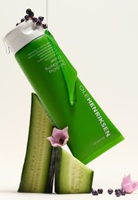 Zielona tubka Ole Henriksen Daily Glow Cleanser z białą nakrętką, ułożona na pokrojonych kawałkach ogórka, podkreślona jagodami i różową kwiatuszką.
