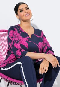 Lächelnde Frau, die auf einem rosafarbenen Stuhl sitzt, trägt marineblaue Hose mit weißen Streifen und ein marineblaues Oberteil mit einem leuchtend pinken Blumenmuster.