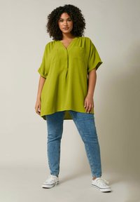 Tunic verde lime con maniche corte e due tasche sul petto, abbinata a jeans azzurri chiari e sneakers bianche. Tessuto liscio, vestibilità comoda.